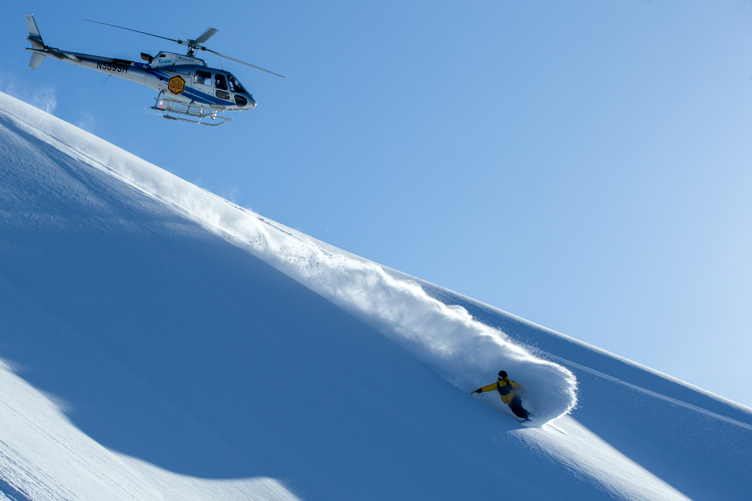 a snowboarder deep in a pow turn with a helicopter flying overhead at Chugach Powder Guides